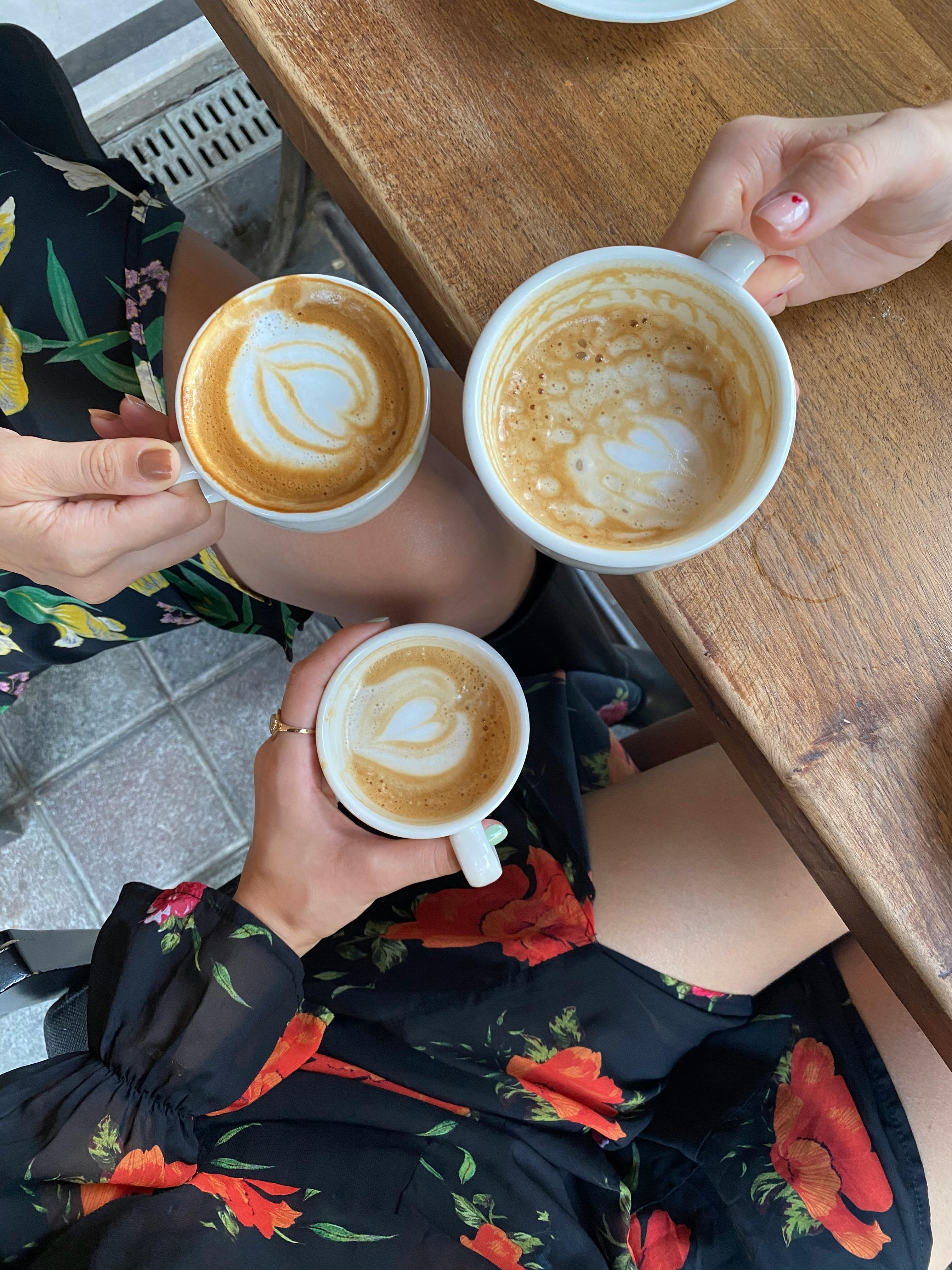 Coffee Time With Friends Three coffee cups held by hands, with intricate latte art on top, placed on a wooden table.