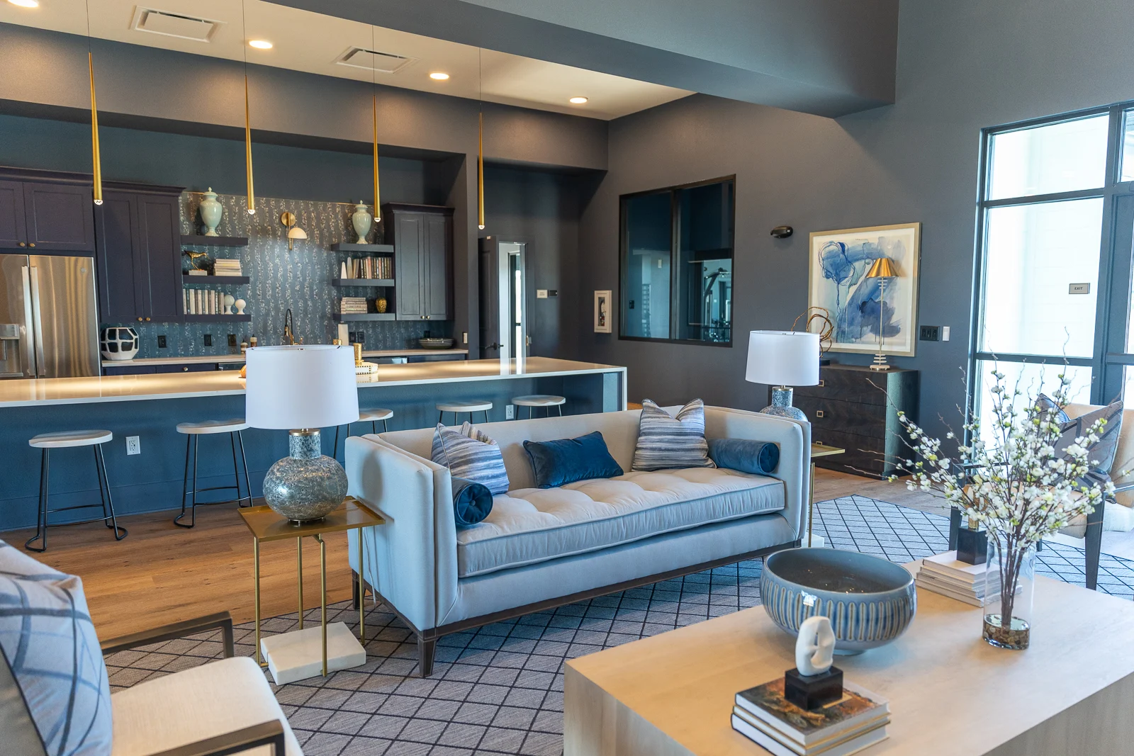 A stylish modern living room featuring a light-colored sofa with blue cushions, a wooden coffee table, and decorative items like books and a small sculpture. The space includes a contemporary kitchen with bar stools, illuminated by pendant lights.