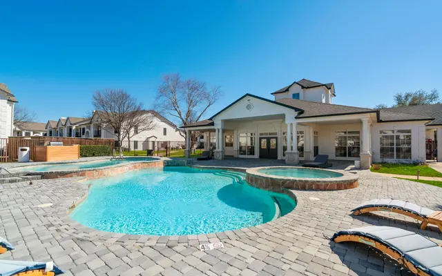 A luxurious pool area at a residential community featuring a large blue swimming pool, sun loungers, a nearby clubhouse, and clear blue skies.