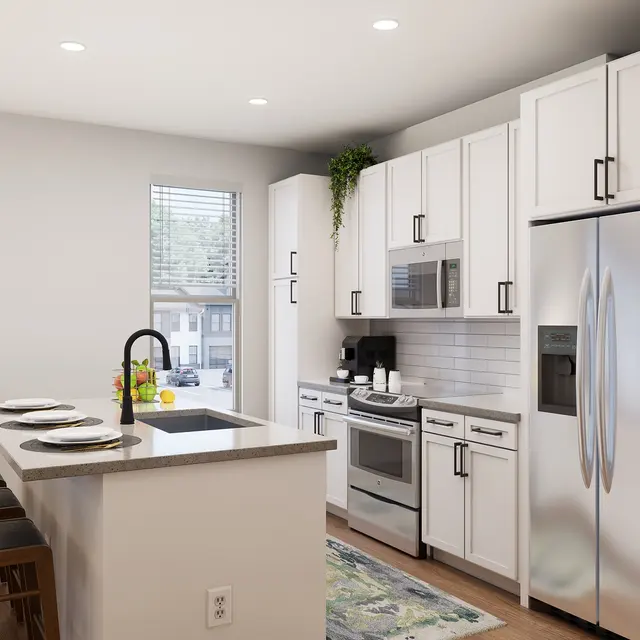 A modern kitchen featuring white cabinetry, stainless steel appliances, and an island with three stools. A window offers natural light, and decorative elements include a rug and a small table with a lamp.