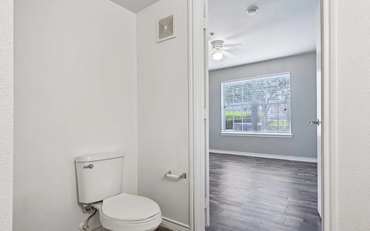 A small bathroom space with a toilet visible on the left and an open doorway leading to a bright room with a window in the background.