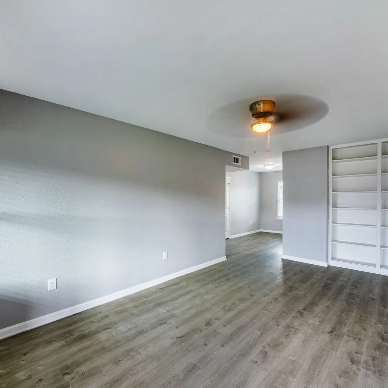 A spacious and modern living room with a light gray wall color and wooden flooring. There's a ceiling fan in the center and a built-in white shelf on the right side. The room is well-lit by natural light coming through the window.