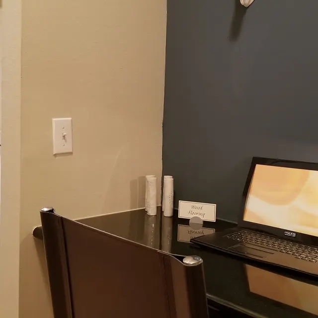 A small home office setup featuring a desk with a laptop, a lamp, and some stationery items. There is a mirror on the wall and the background shows a bathroom door and towels.