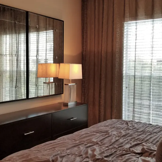 A cozy bedroom featuring a brown bedspread on a neatly made bed, a modern dresser with a large mirror above, and a lamp. The window is covered with sheer curtains, allowing light to filter through, and the room has a warm and inviting atmosphere.