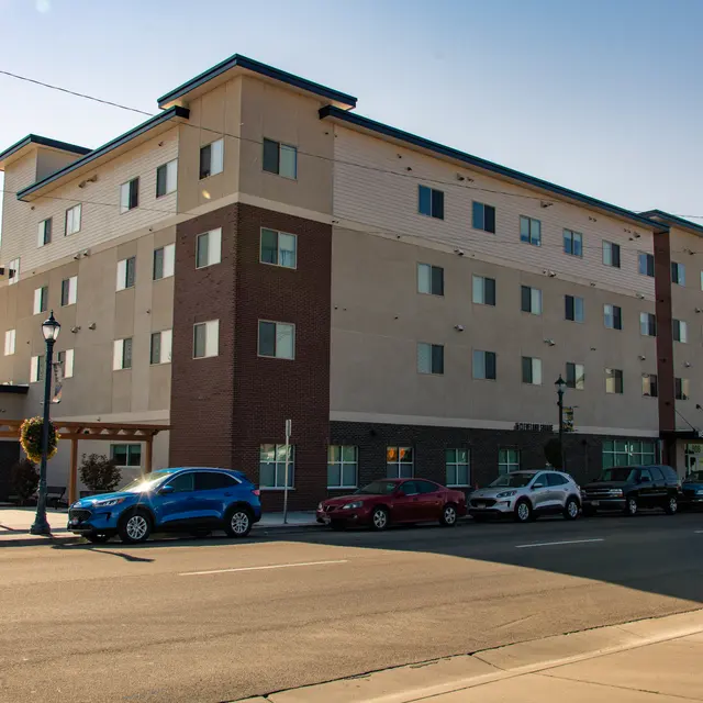 Modern Commercial Building Exterior A modern four-story building with a mix of light-colored and dark brick exterior. Several parked cars line the street in front of the building on a clear day.