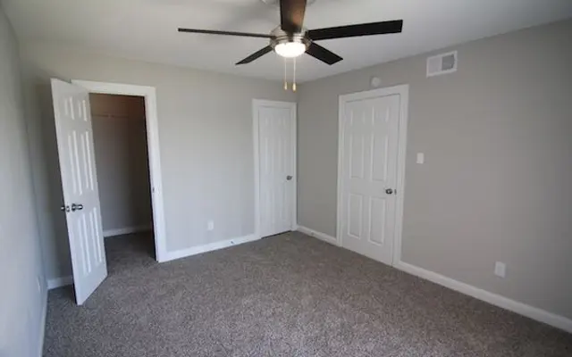 Empty Room Interior A spacious, empty room with beige carpet and light grey walls. The room has a ceiling fan and two closed doors leading to closets.