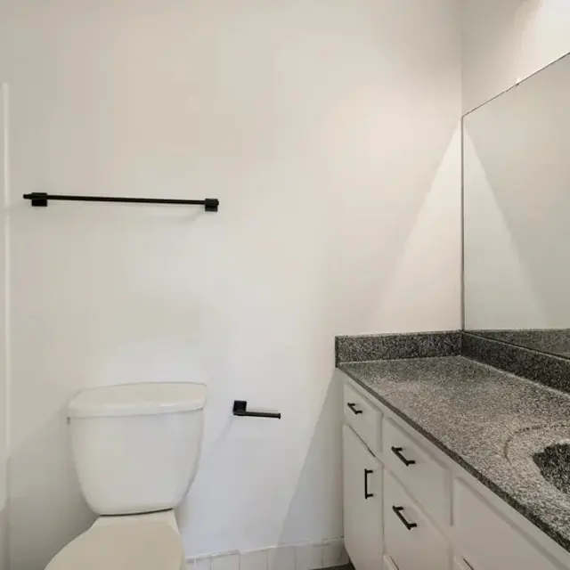 A modern bathroom featuring a white bathtub and toilet, a grey granite countertop with a sink, and a large mirror. There are minimalistic black fixtures and a towel rack on the wall.