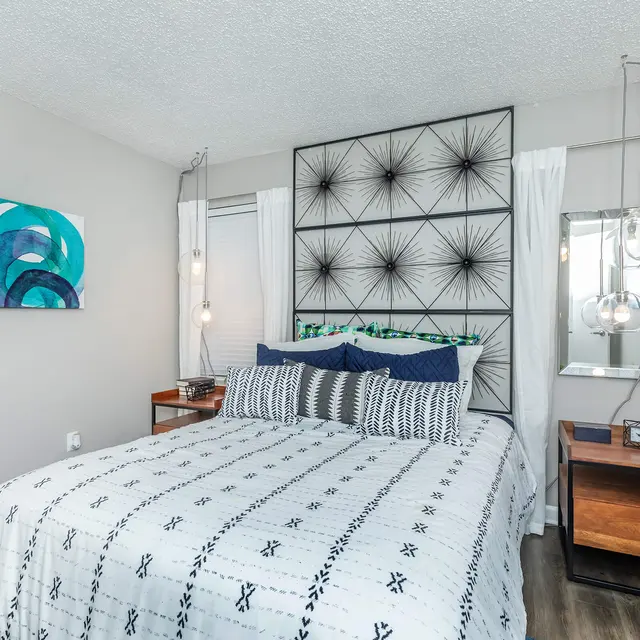 A modern bedroom featuring a large bed with patterned bedding and two decorative pillows. There's a geometric headboard, side tables with lamps, and abstract wall art in shades of blue and green. The room has light-colored walls and wood flooring.