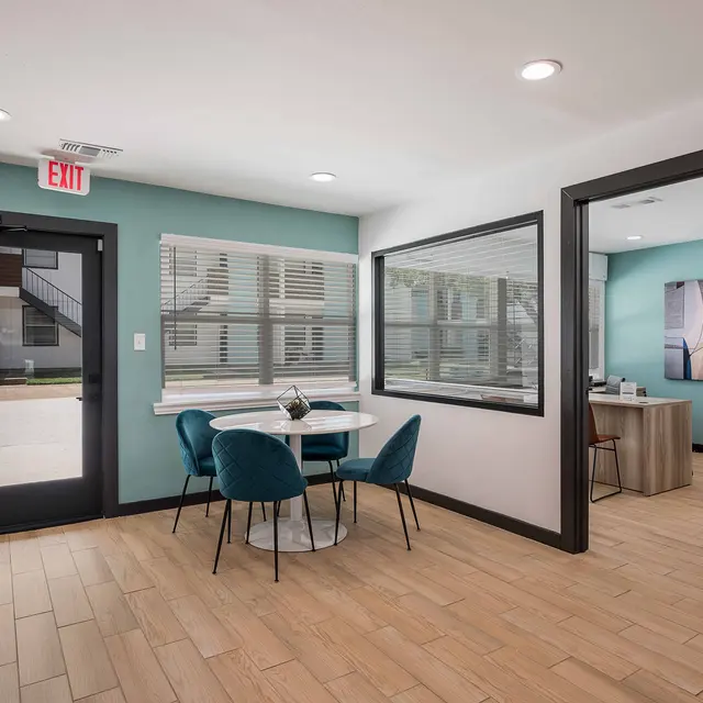 A modern interior space featuring a small dining table with teal chairs, light wood flooring, and large windows. There is an exit door on the left and a reception desk visible in the background.