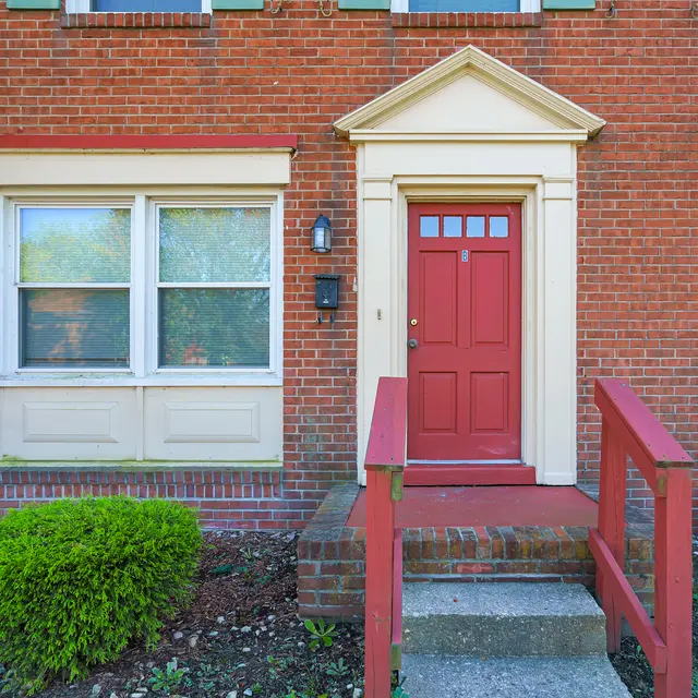 Front entrance of a brick building with a red door and small steps leading to it, flanked by two sets of windows