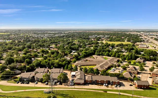 Aerial view of a suburban area with numerous trees, buildings, and roads. The landscape includes green lawns and parking areas.