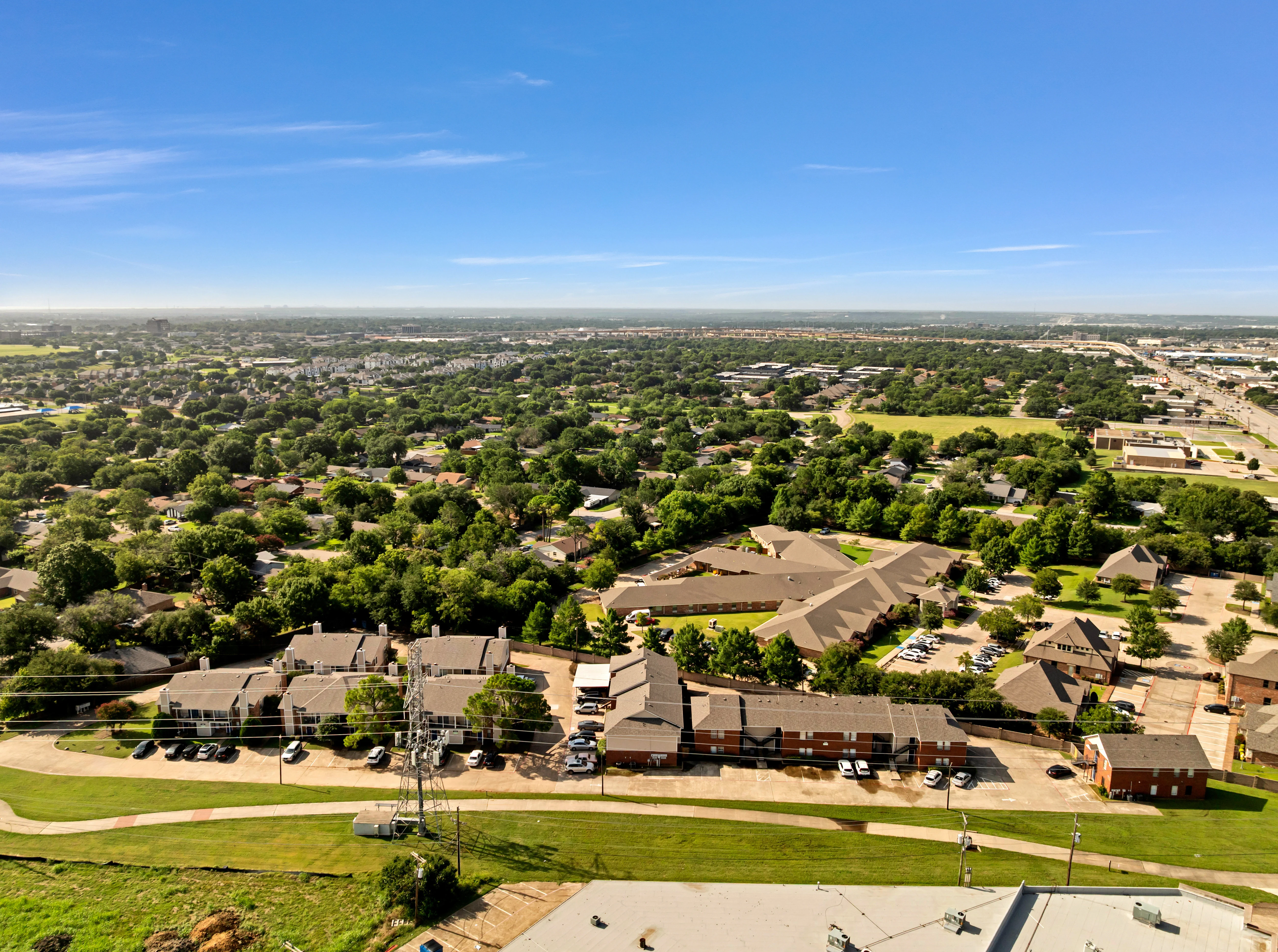 Aerial view of a suburban area with numerous trees, buildings, and roads. The landscape includes green lawns and parking areas.