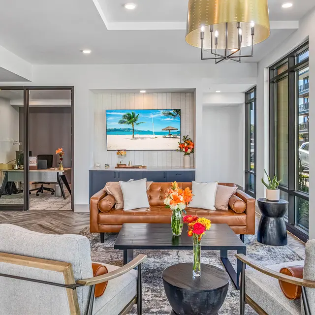 A modern lobby area featuring a comfortable leather sofa, two armchairs, a coffee table with fresh flowers, and a painting of a beach scene on the wall. Large windows provide natural light.