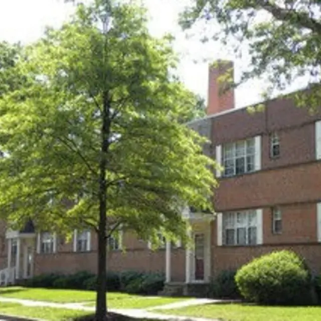 A brick apartment building with several windows, surrounded by green trees and grass.