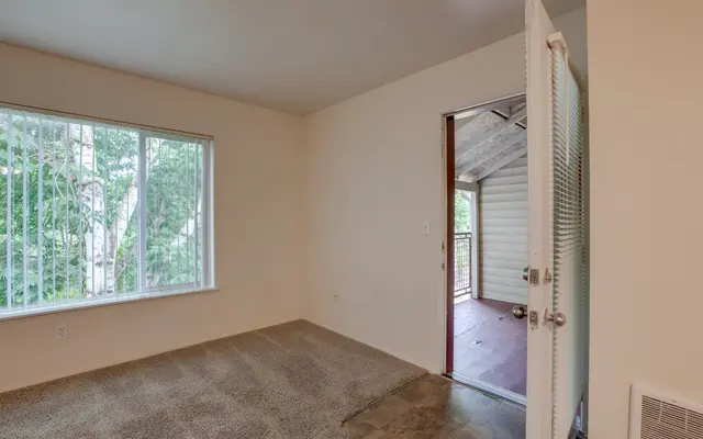 An empty room with a light-colored wall and a window showing greenery outside. A door is open, leading to another space with a covered porch.