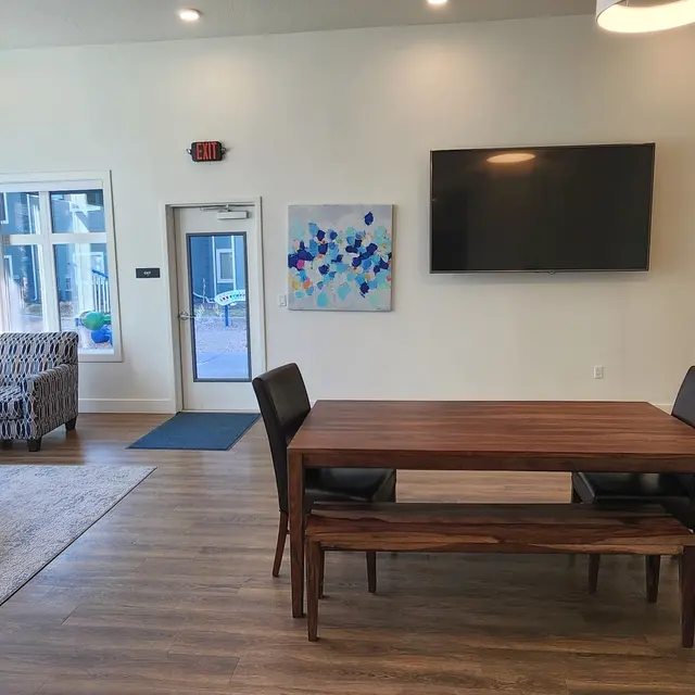 Canyon Terrace A modern communal living area featuring a comfortable sofa, a coffee table, and a dining table with chairs. There is a large TV mounted on the wall and various artworks displayed. Large windows let in natural light, creating an inviting atmosphere.