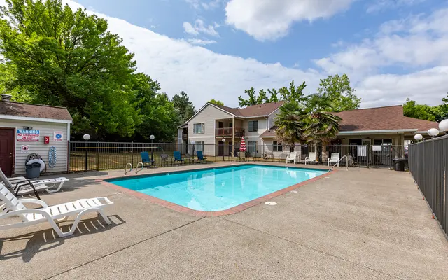 A sunny pool area with a clear blue pool surrounded by lounge chairs and a fence, set against a backdrop of trees and a two-story building.