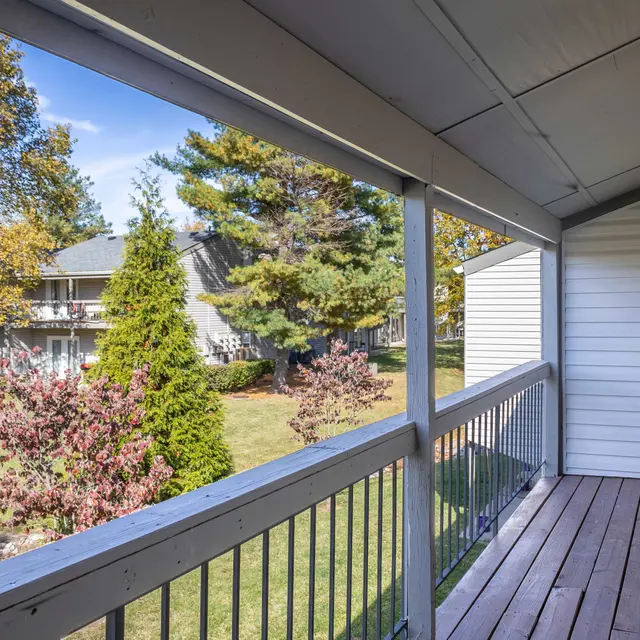 Balcony View in Autumn A balcony view overlooking a green lawn with some trees and autumn foliage. There are neighboring buildings in the background.