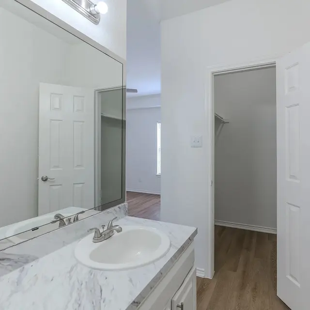 A modern bathroom with a marble countertop, a large mirror above the sink, white cabinetry, and a small closet visible through a door to the right.