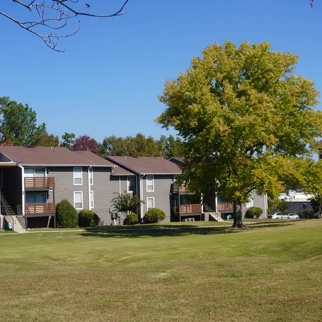 A view of an apartment complex with multiple buildings surrounded by green grass and trees, under a clear blue sky.