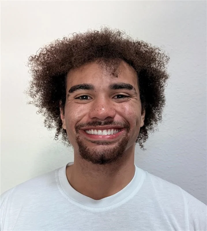 Smiling Young Man with Afro Hairstyle A young man with curly, afro-style hair and a goatee, smiling brightly at the camera while wearing a plain white t-shirt.