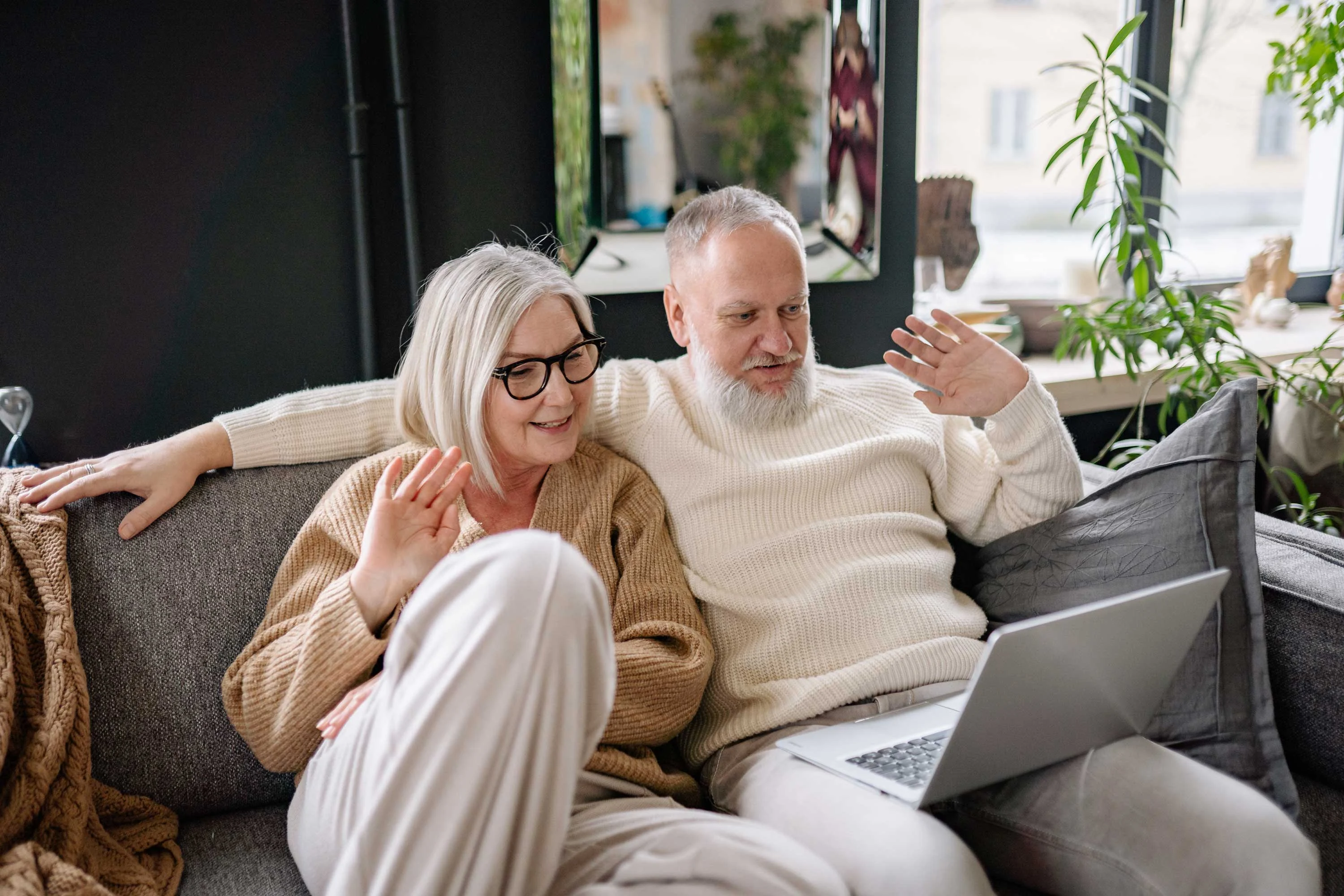 Elderly Couple on a Video Call An elderly couple sitting comfortably on a couch, happily waving at a laptop during a video call. They are wearing cozy sweaters and relaxed pants, surrounded by a modern living room with plants and decorative items.