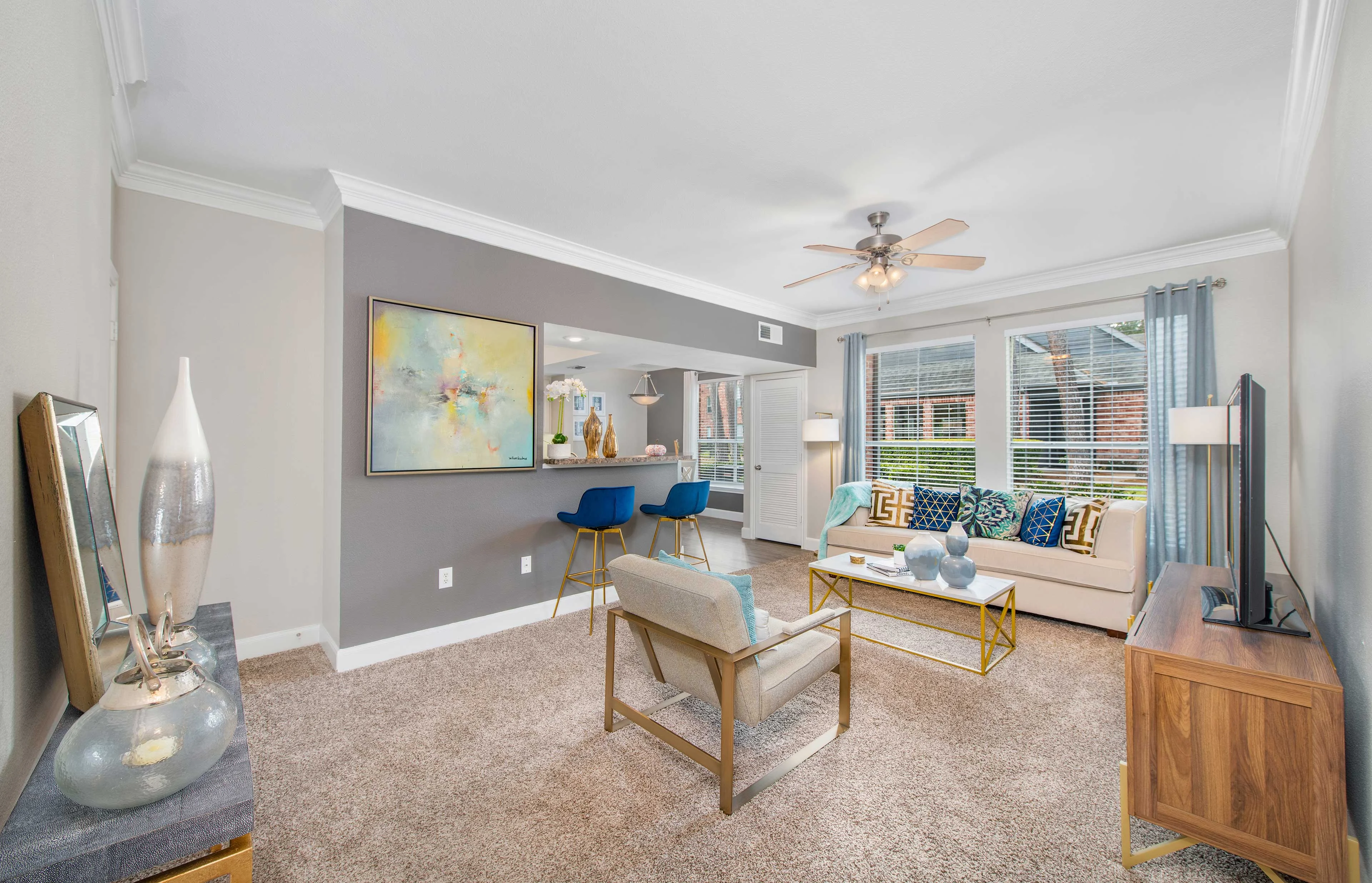 Stylish Modern Living Room A modern living room with a beige sofa, colorful decorative cushions, and a stylish coffee table. The room has large windows letting in natural light, a ceiling fan, and a light-colored wall with artwork and a mirror. There's a side table and a sleek television stand.