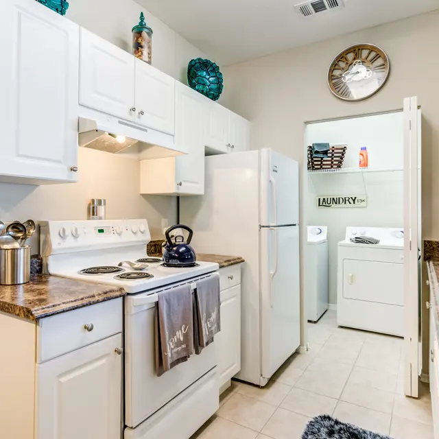 A modern kitchen with white cabinetry and granite countertops, featuring a stove, kettle, and refrigerator. A laundry room is visible through an open door in the background.