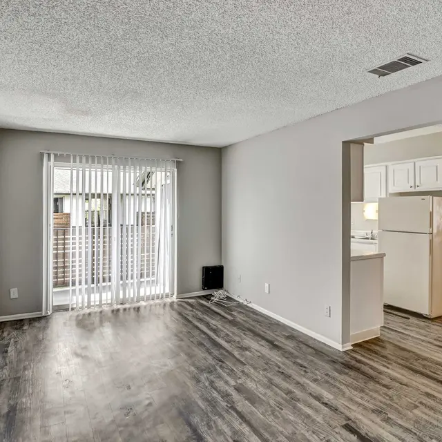A spacious and empty apartment interior featuring a living area with light wood flooring, vertical blinds on a large window, and a view into a kitchen area with white cabinetry and a refrigerator visible.