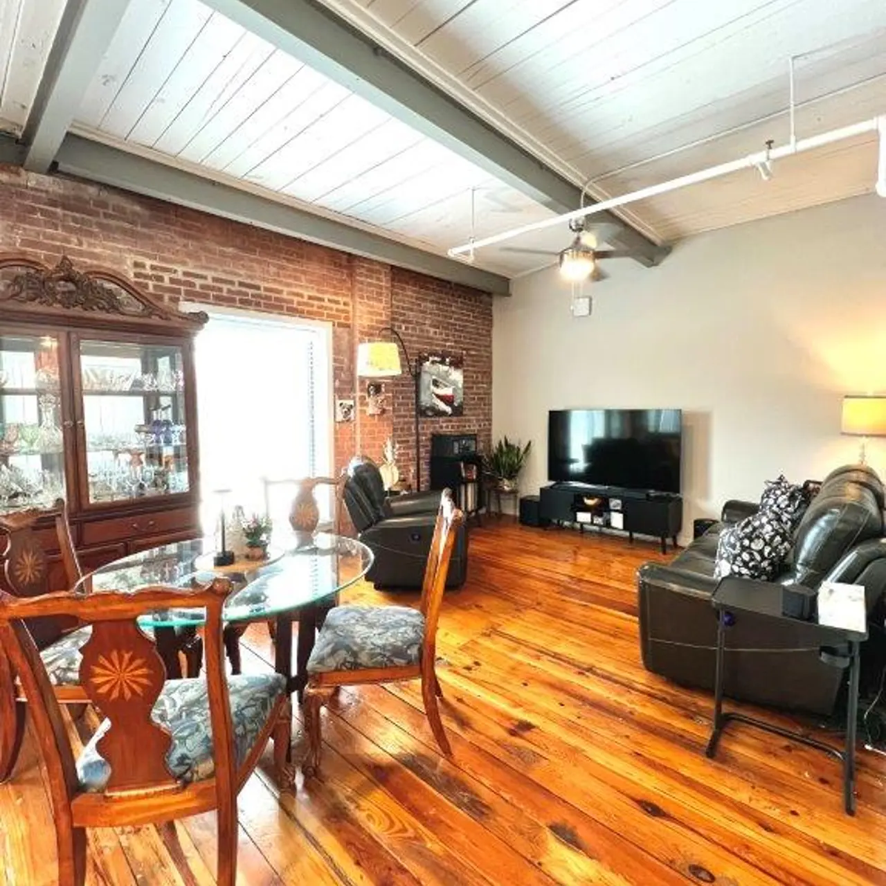 A cozy living room featuring a wooden floor, exposed brick wall, and ceiling beams. There is a glass dining table with ornate chairs and a decorative cabinet. A television is against the wall with two leather armchairs nearby.