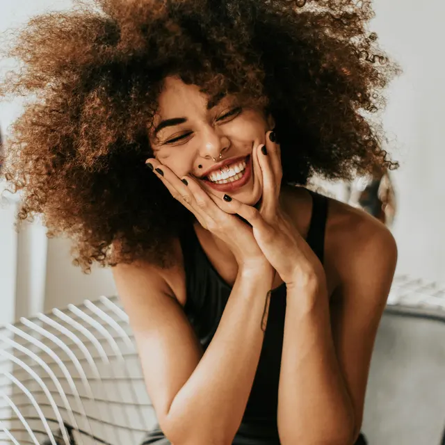 Joyful Woman with Curly Hair A woman with voluminous curly hair, smiling broadly while sitting on a modern chair. She appears joyful and stylish, with her hands on her cheeks.