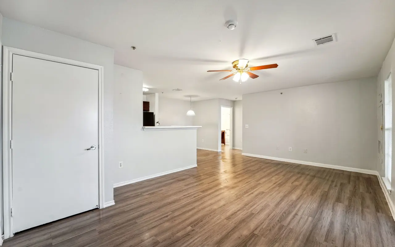 A spacious living room with light wooden flooring, a ceiling fan, and an open layout leading to a kitchen area.