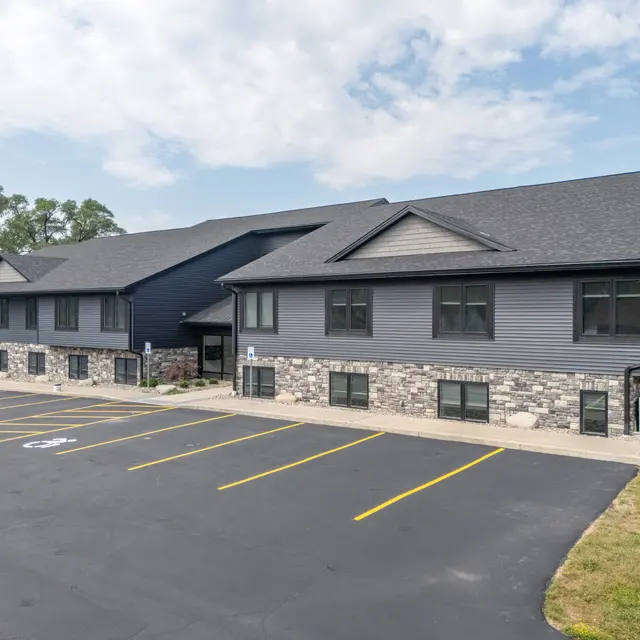 A modern two-story building with a combination of dark siding and stone exterior. There is a paved parking lot with several empty spaces in the foreground and green grass surrounding the structure.