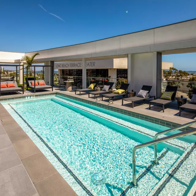 Rooftop Pool Area A rooftop pool area featuring a modern design with a swimming pool, lounge chairs, and palm trees. The backdrop shows a clear blue sky and a distant cityscape.