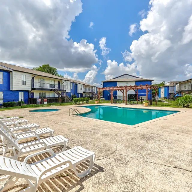A view of a pool area surrounded by apartment buildings painted in blue. The pool is clear, with lounge chairs lined up beside it. In the background, there are cloudy skies and green grass surrounding the area.