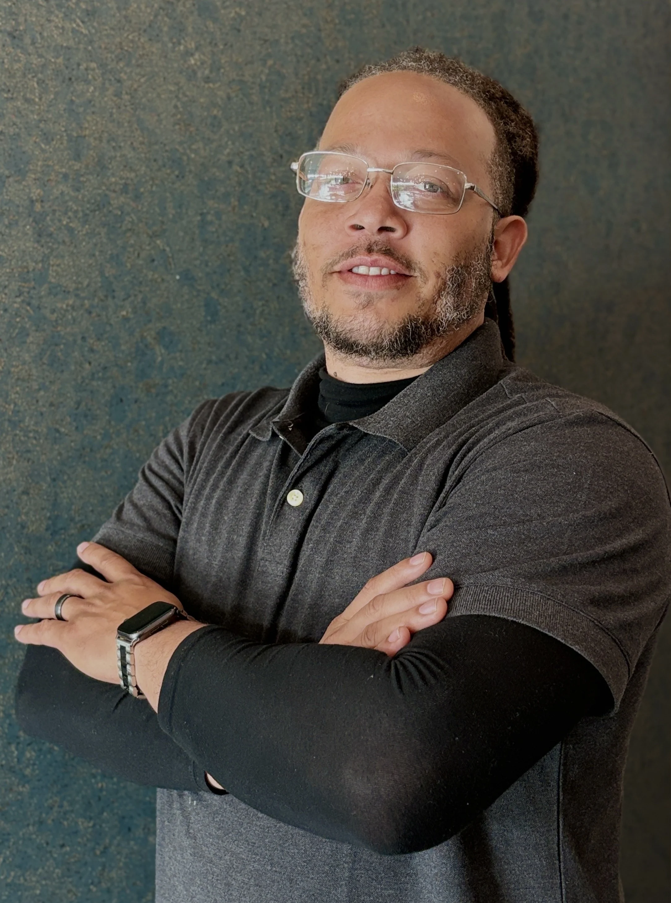 Confident Man with Glasses A man with glasses and dreadlocks standing with arms crossed, wearing a dark polo shirt and black long-sleeve undershirt, smiling against a textured blue background.