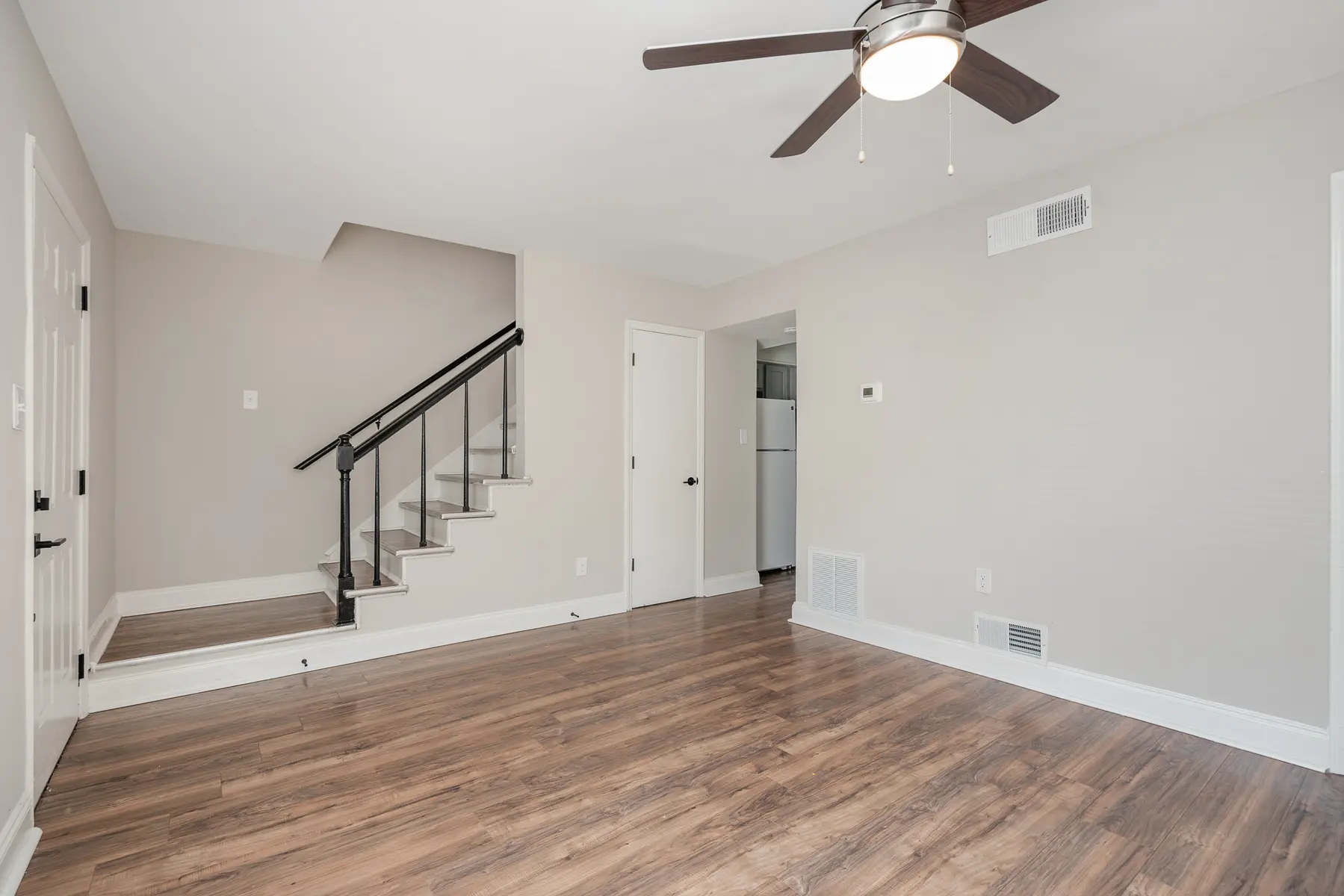 A modern living room with light brown wooden flooring, a ceiling fan, and a staircase leading to another level. The walls are painted in a neutral shade, and there is a door leading to a kitchen area in the background.