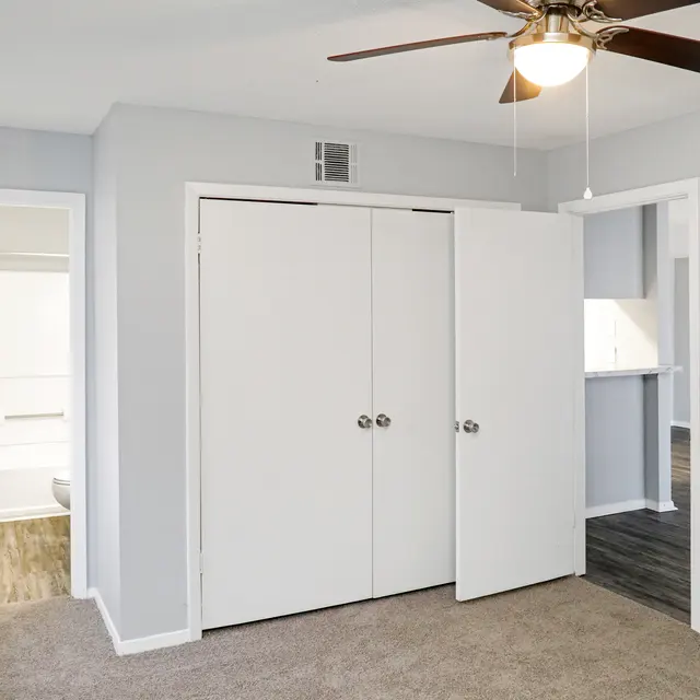 A view of a light gray bedroom featuring a ceiling fan, with two doors leading to other rooms. To the left is a bathroom and to the right an adjacent room. The floor is carpeted and there is a small closet with double doors.
