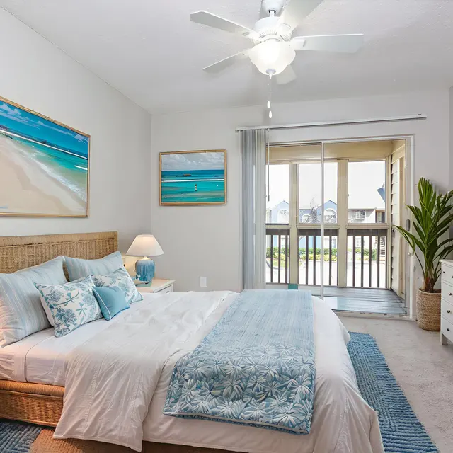 A serene beach-themed bedroom featuring a light color palette with blue accents. The room has a queen-sized bed with decorative pillows and a bedspread, a beige bed frame, and two bedside tables with lamps. There are abstract ocean-themed paintings on the walls, a large window with sliding doors leading to a balcony, and a TV mounted on the wall. A plant and a white dresser are also present, adding to the relaxed atmosphere of the space.