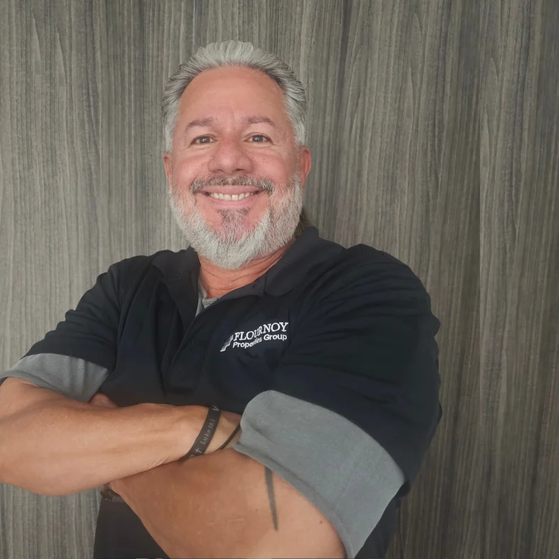 Smiling Man in Business Casual Attire A smiling man with grey hair and a beard, crossing his arms in front of a wooden textured background. He is wearing a black polo shirt with grey sleeves.