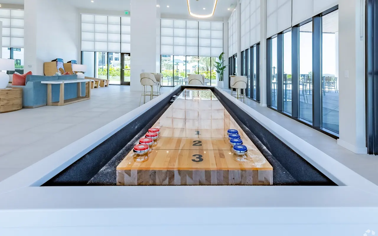 A sleek indoor shuffleboard setup with a wooden playing surface, featuring numbered scoring sections and colored pucks on either end. The room is bright and modern, with large windows and minimalist decor.