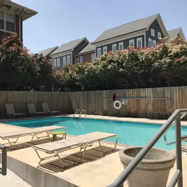 A peaceful swimming pool area surrounded by a wooden fence, featuring lounge chairs and a few trees. The water is calm and the sky is clear and blue.