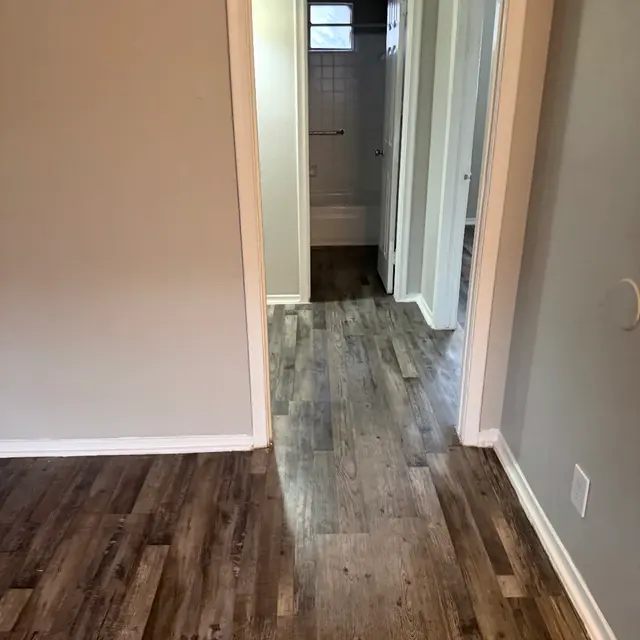 A view of a hallway in a residential space with dark wood-like flooring and light gray walls. A bathroom door is visible to the left, and there are additional doorways leading to other rooms.