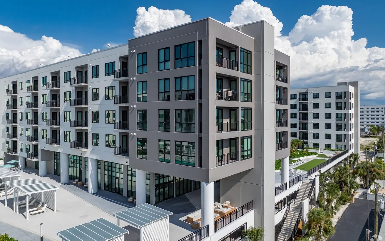 A modern apartment building with multiple floors and large windows, featuring a sleek, contemporary design and surrounded by greenery and outdoor seating areas.
