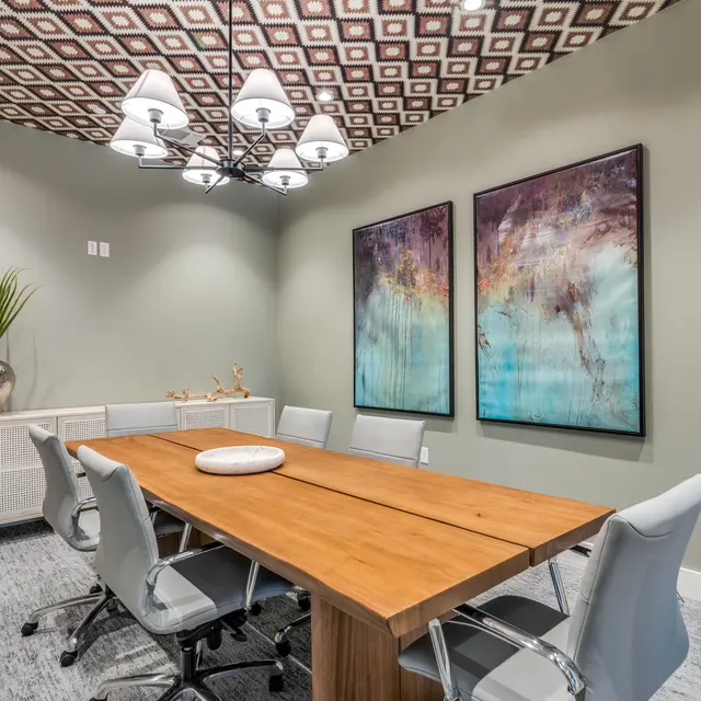 A modern meeting room featuring a long wooden table surrounded by six light gray chairs. Two large decorative vases with green plants are positioned in the corner. The walls are painted in a soft green, with two colorful abstract art pieces hung. The ceiling is adorned with a geometric design and a stylish light fixture hangs above the table.