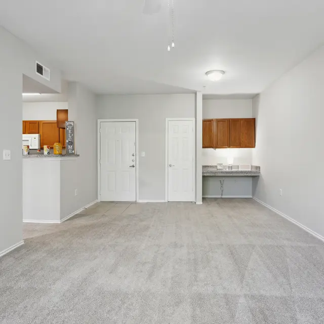A spacious, empty living room with light gray walls and carpeting, featuring a small kitchenette visible in the background.