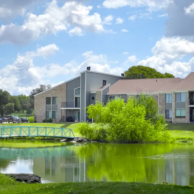 A serene scene featuring a pond, a green bridge, and landscaped apartments surrounded by lush trees under a cloudy sky.