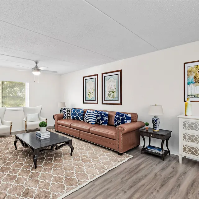 A modern living room featuring a brown sofa with blue patterned pillows, a coffee table in the center, and two white armchairs facing a window. Wall art is displayed, and a light hardwood floor is visible.