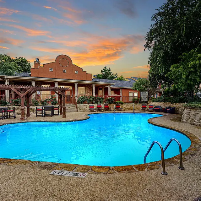 A beautifully lit swimming pool at dusk, surrounded by lounge chairs and trees, with a building in the background under a colorful sky.