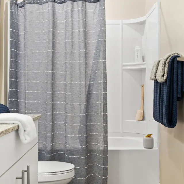 A modern bathroom featuring a shower with a gray curtain, a white toilet, and wooden flooring. There is a shower shelf with a few items and a blue towel hanging nearby.